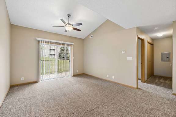 Spacious apartment living room with carpeted flooring, a vaulted ceiling, a ceiling fan, and a large sliding glass door with vertical blinds providing natural light and access to a private patio or balcony.