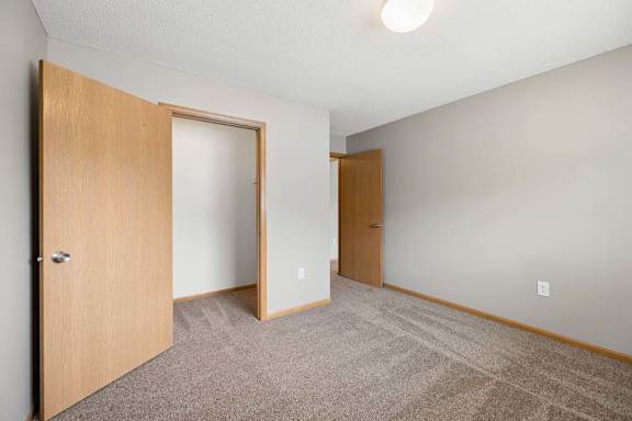 Bright apartment bedroom with carpeted flooring, an open closet with sliding doors, neutral walls, and a separate entry door for convenient access.