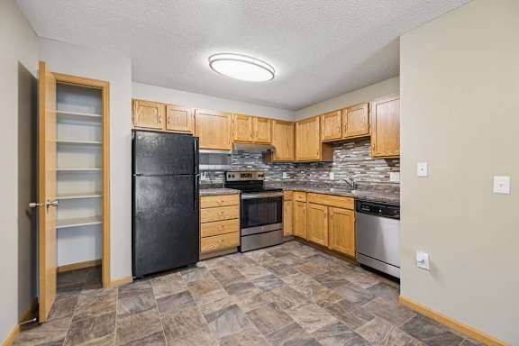 Apartment kitchen with wood cabinetry, a tile backsplash, laminate countertops, black and stainless steel appliances, and a pantry with built-in shelving in a functional U-shaped layout.