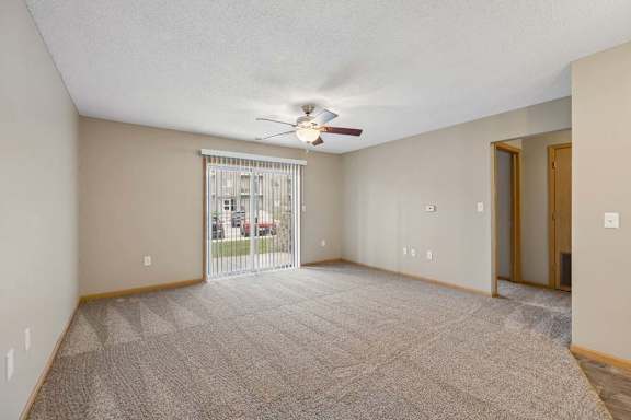 Spacious apartment living room with carpeted flooring, a ceiling fan, and a large sliding glass door with vertical blinds providing natural light and access to a private patio or balcony.