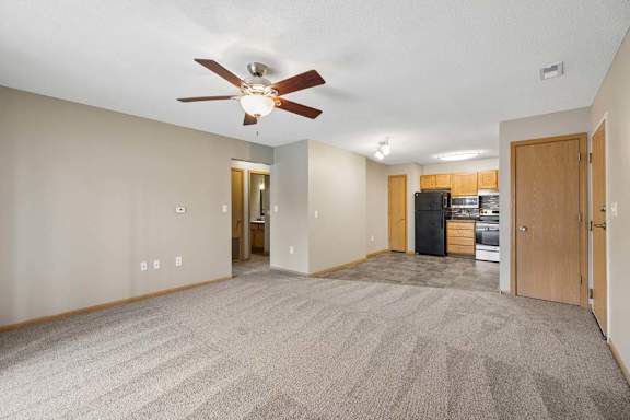 Spacious open-concept apartment living room with carpeted flooring, a ceiling fan, and a view into the kitchen featuring wood cabinetry, black appliances, and an adjacent dining area.