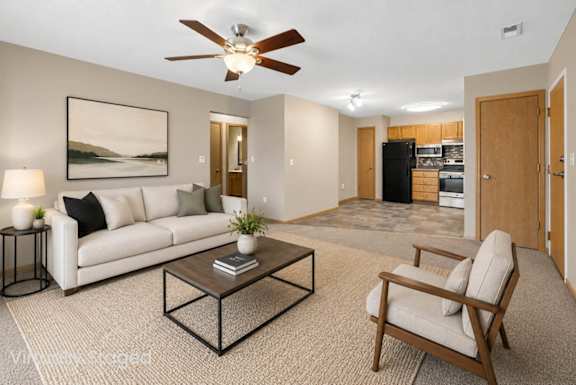 Virtually staged living area with a sofa, coffee table, accent chair, ceiling fan, and an open connection to the kitchen.