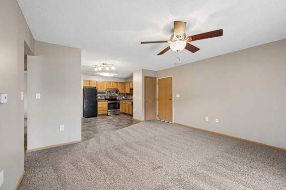 Spacious open-concept apartment living room with carpeted flooring, a ceiling fan, and a view into the kitchen featuring wood cabinetry, black appliances, and an adjacent dining area.