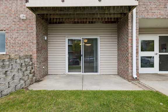 Covered ground-floor apartment patio with a concrete surface, a large sliding glass door, brick and siding exterior, and direct access to a landscaped lawn.