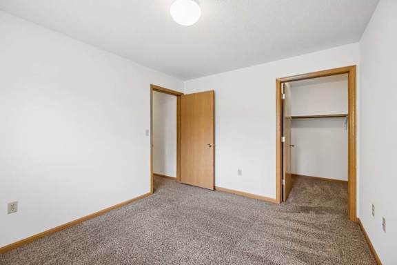 Bright apartment bedroom with carpeted flooring, neutral walls, an open closet with a hanging rod, and a separate walk-in closet for additional storage.