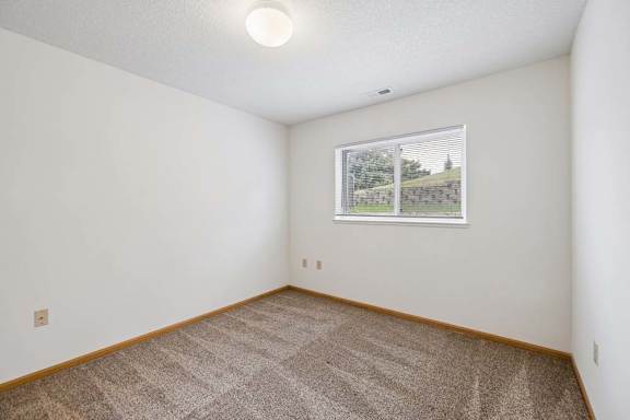 Bright apartment bedroom with carpeted flooring, neutral walls, and a window providing natural light with views of landscaped green space.