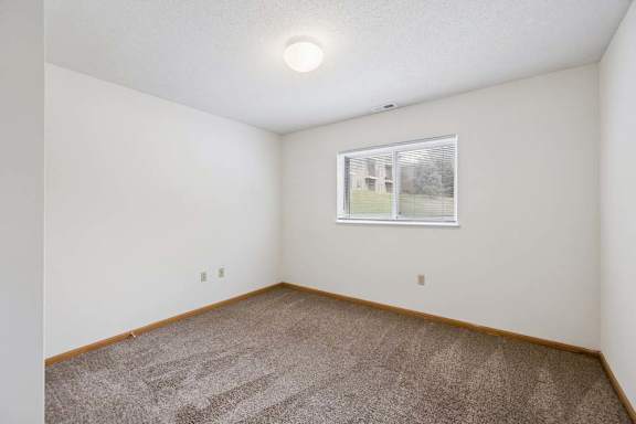 Spacious apartment living room with carpeted flooring, a ceiling fan, and a large sliding glass door with vertical blinds providing natural light and access to a private patio or balcony.