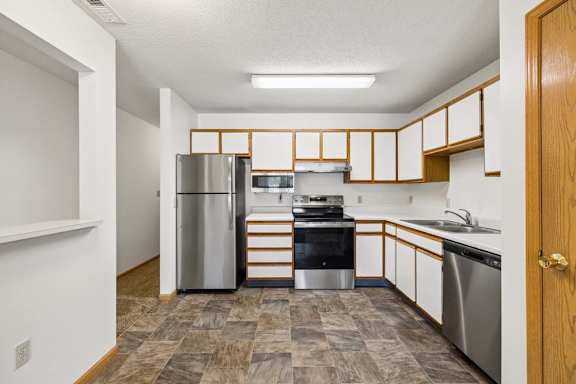 Apartment kitchen with white and wood-tone cabinetry, stainless steel refrigerator and dishwasher, black oven, laminate countertops, and a functional U-shaped layout with a pass-through opening.