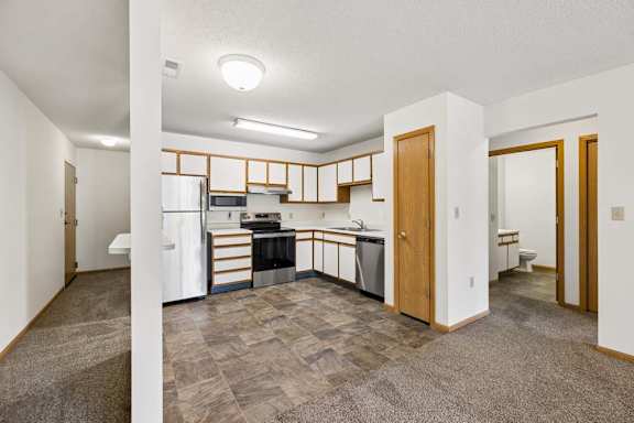 Open-concept apartment with a kitchen featuring white and wood-tone cabinetry, stainless steel and black appliances, a pantry, and adjacent carpeted living and dining areas.