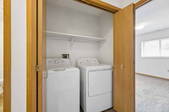 Apartment laundry closet with side-by-side washer and dryer, a built-in shelf above, and wood bifold doors for convenient in-home laundry access.