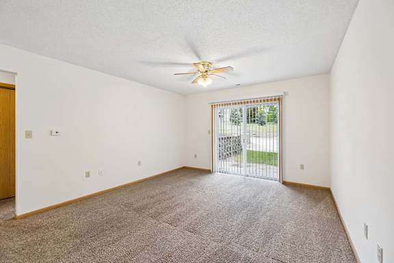 Spacious apartment living room with carpeted flooring, a ceiling fan, and a large sliding glass door with vertical blinds providing natural light and access to a private patio or balcony.