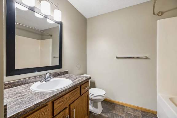 Bathroom vanity with wood cabinetry, stone-look countertop, framed mirror, modern lighting, and a tub-shower combination.