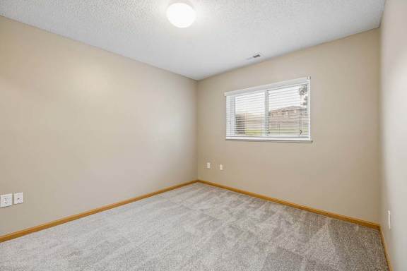 Unfurnished bedroom with wall-to-wall carpeting and a wide window with blinds overlooking the community grounds.