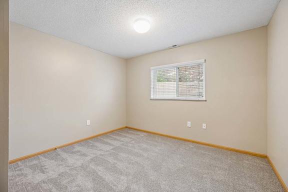 Bedroom with beige walls, plush carpeting, and a window with horizontal blinds providing natural light.