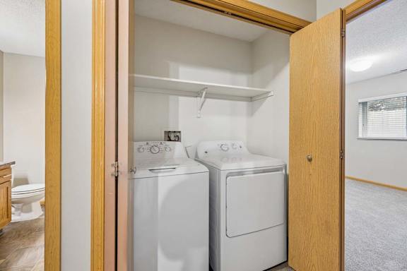 In-home laundry closet with a full-size washer and dryer, overhead shelf, and folding doors between the bedroom and bathroom.