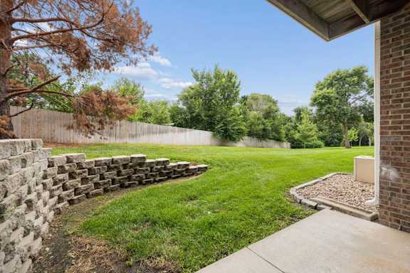 Ground-level patio overlooking a grassy courtyard with mature trees, a wood privacy fence, and stone retaining wall.