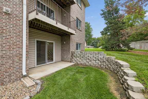 Ground-floor apartment patio with sliding glass doors, brick exterior, curved stone retaining wall, and grassy surroundings.