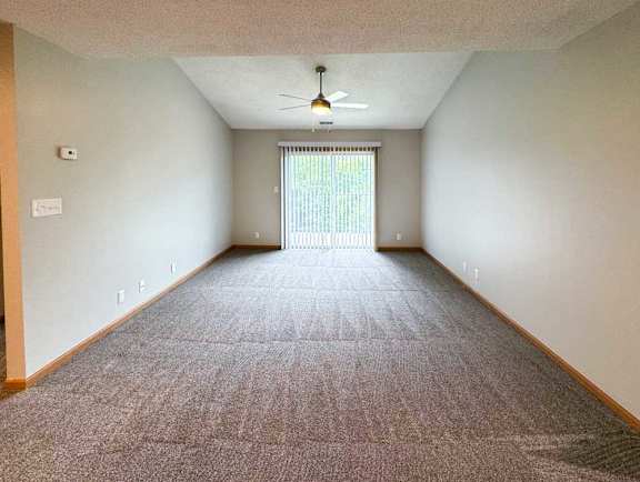 A living room with a carpeted floor, a ceiling fan, and a sliding glass window to a balcony.