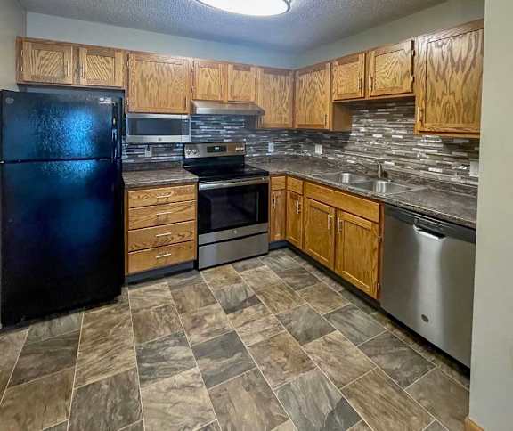 A kitchen with a black refrigerator, wooden cabinets, tile backsplash, stainless steel appliances, and granite countertops.