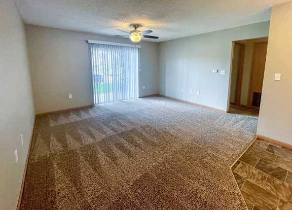 A living room with a carpeted floor and a sliding glass door with blinds.