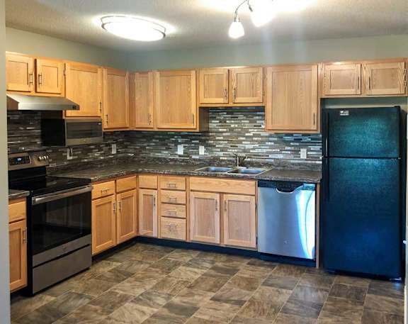 A kitchen with wooden cabinets, a black refrigerator, stainless steel appliances, a tile backsplash, and granite countertops.