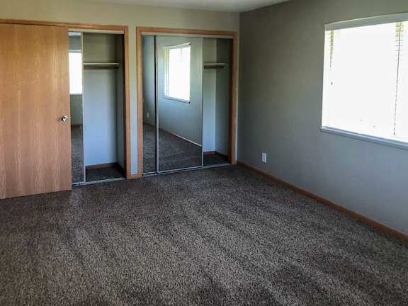 A bedroom with a carpeted floor, two mirrored-door closets, and a large window.