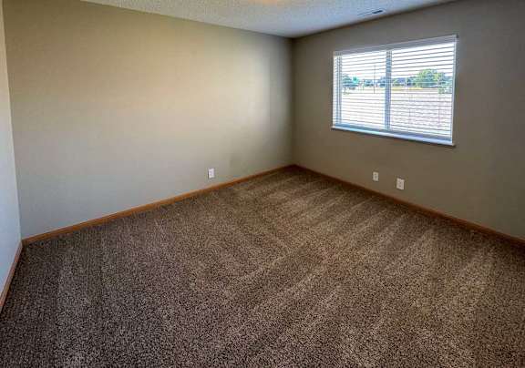 A bedroom with a carpeted floor and a wide window with blinds.