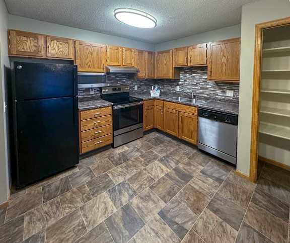 A kitchen with a black refrigerator, wooden cabinets, stainless steel appliances, a tile backsplash, and granite countertop
