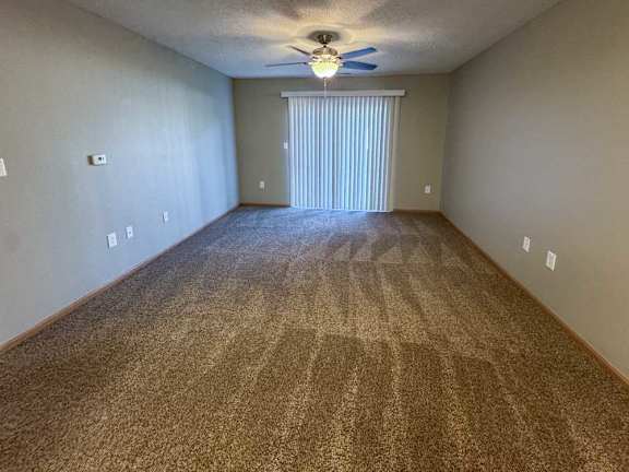 A living room with a carpeted floor, ceiling fan, and a sliding glass door with vertical blinds.