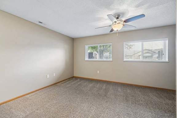Spacious apartment bedroom with carpeted flooring, a ceiling fan, and two windows with blinds providing natural light and neighborhood views.
