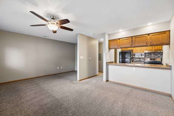 Open-concept apartment living room with carpeted flooring, a ceiling fan, and a breakfast bar overlooking the kitchen with wood cabinetry and black appliances
