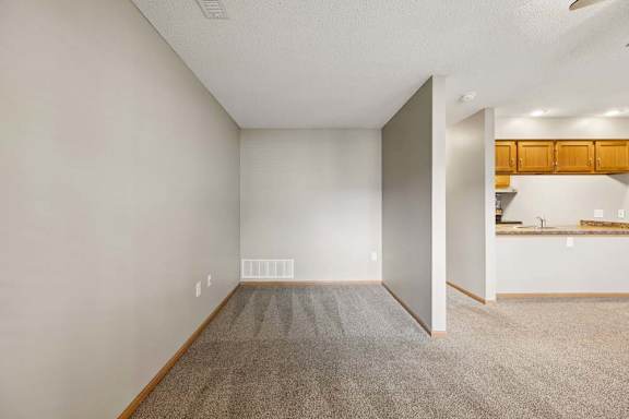 Spacious apartment living area with carpeted flooring and a flexible nook, adjacent to an open kitchen with a breakfast bar and wood cabinetry.