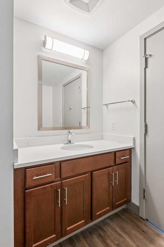 A bathroom with wooden cabinets, a white granite vanity sink, and a framed mirror with overhead lighting.