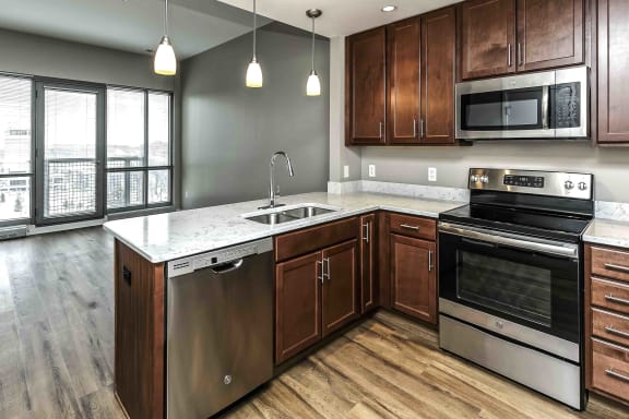 View from inside the kitchen, looking at the the dishwasher, sink, oven and microwave. Living room of a one bedroom apartment at Midtown Crossing is seen in the background.