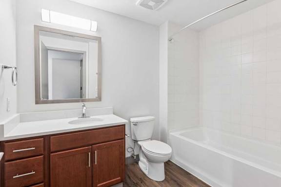 A bathroom with wooden cabinets, white granite counters, a mirror with overhead lighting, and a tub shower.