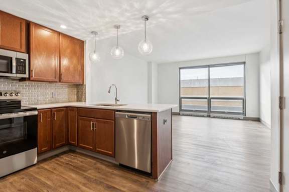 A kitchen with wooden cabinets and a stainless steel dishwasher, in front of a living room with a large window.