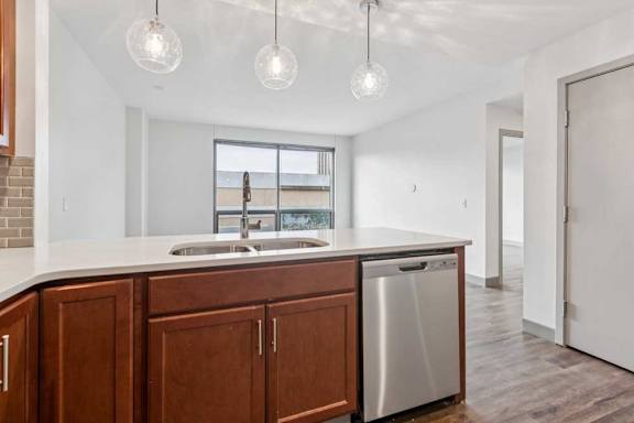 A kitchen peninsula with wooden cabinets and stainless steel dishwasher, in front of a living room.