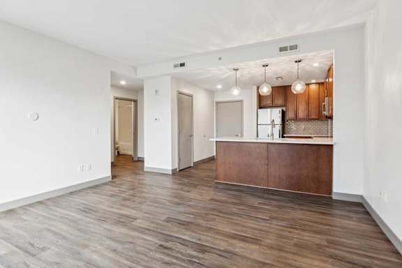 A spacious living room with hardwood-style flooring, open to a kitchen and hallway leading to a bedroom.