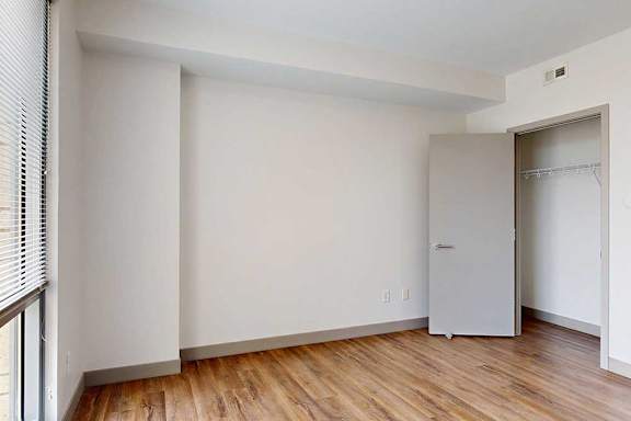 A bedroom with hardwood-style flooring, a wide walk-in closet, and a floor-to-ceiling window.