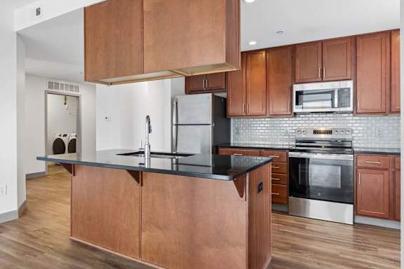 Apartment kitchen with wood cabinetry, black countertops, stainless steel appliances, tile backsplash, and visible laundry area.