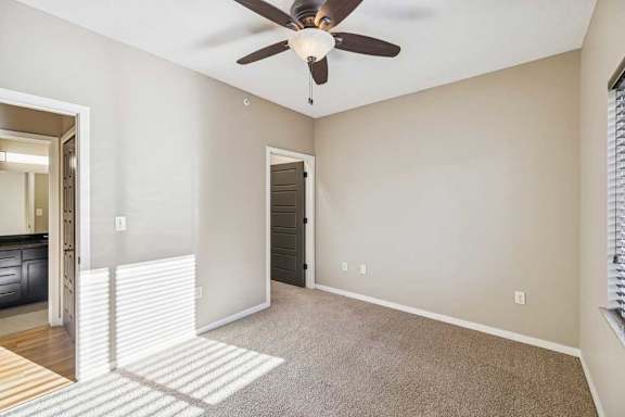 A carpeted bedroom with a ceiling fan. The bedroom door is on the left and the bathroom is in the background. The closet door is on the right.
