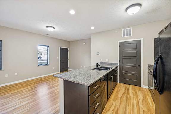 A kitchen with grey granite countertops, black appliances, and a large kitchen island. There is a large living room with wooden floors throughout. There is a window next to the front door in the living room.