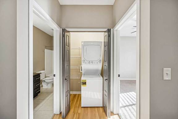 The end of a hallway with the bathroom on the left, the stacked washing machine straight ahead, and a carpeted bedroom on the right