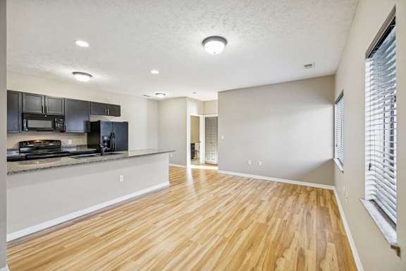 A large living room with wooden floors and a large kitchen island. Black appliances are in the kitchen. There is a hallway in the background.