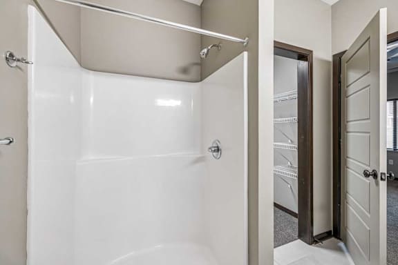 Bathroom with white tub and shower surround, chrome fixtures, and neutral walls, with an adjacent walk-in closet featuring wire shelving and a paneled door in this apartment interior.