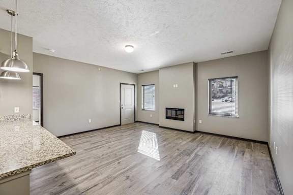 Open-concept apartment living room with wood-style flooring, neutral walls, and large windows with blinds, featuring a wall-mounted fireplace and granite breakfast bar with pendant lighting.