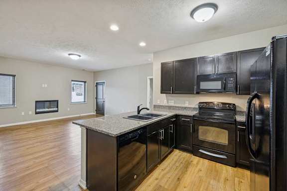A kitchen with dark wooden cabinets, granite countertops, black appliances, and a peninsula countertop in front of a spacious living room.