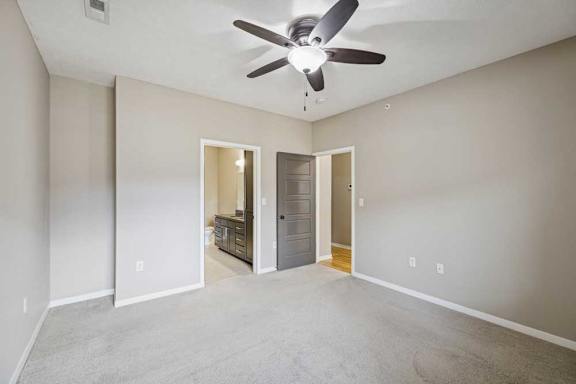 A carpeted bedroom with an attached bathroom, a ceiling fan, and a door open to the living room.