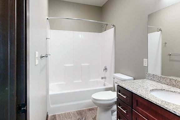 A hallway bathroom with dark wooden cabinets, light granite counter, and a tub shower with chrome finishes.
