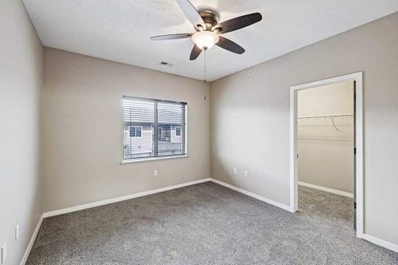 Apartment bedroom featuring neutral carpeting, a ceiling fan with light, and a window overlooking the exterior.